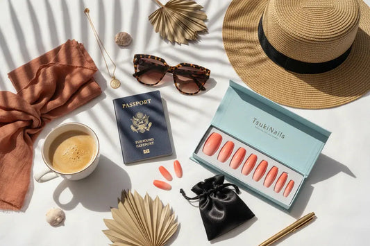 Chic travel flat lay with Tsuki Nails coral ombre press on nails, passport, sunglasses, straw hat on white bed.