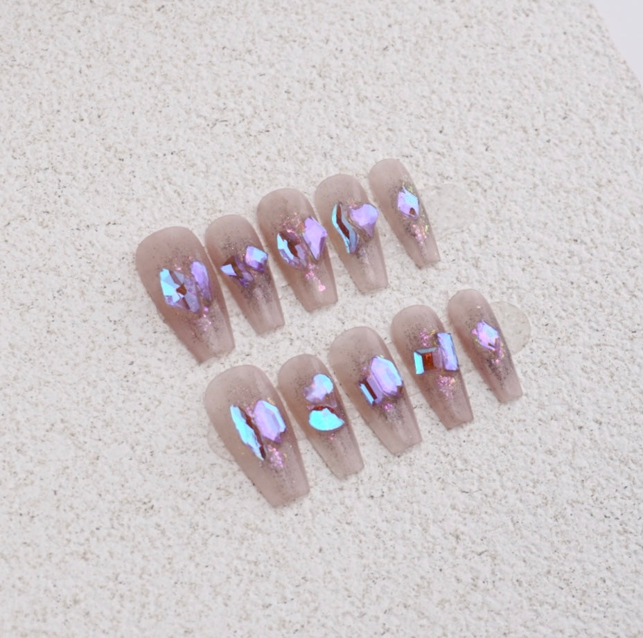 Long coffin press-on nails featuring large iridescent purple crystal shards and glitter on a sheer smokey nude base.


