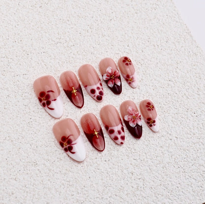 Almond shaped press-on nails featuring burgundy red flowers, white french tips, blush aura design, and gold cross charm.

