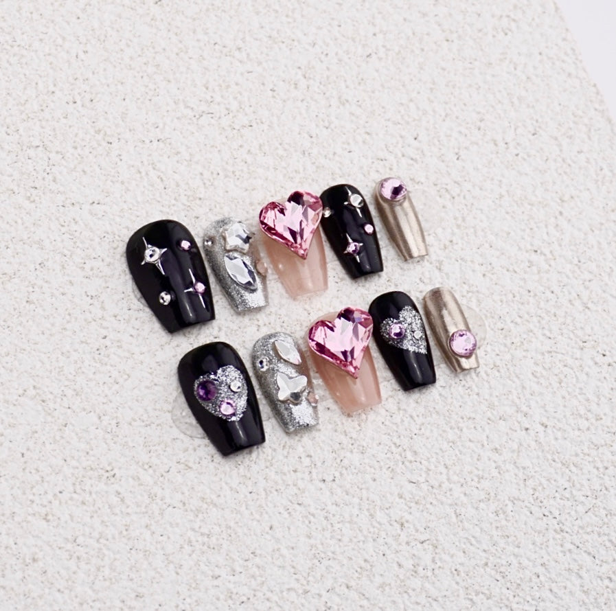 Edgy black and silver press-on nails featuring a large pink heart crystal, glitter accents, and metallic chrome finish.

