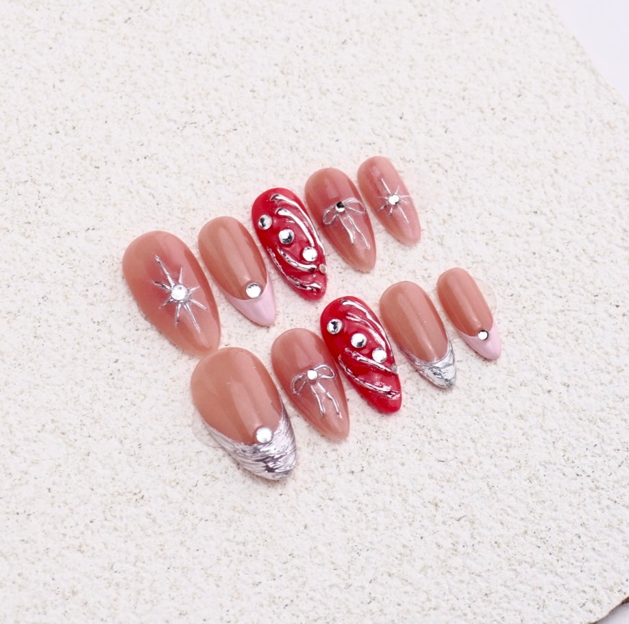 Almond press-on nails featuring 3D silver bows, stars, red chrome liquid swirls, and french tips.

