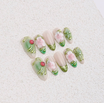 Almond press-on nails featuring green chrome vines, pink 3D flowers, and a red ladybug charm on a nude base.

