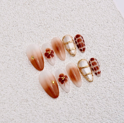 Almond press-on nails featuring brown vertical gradient, vintage four-petal flower, textured gold chrome lines, and polka dots.

