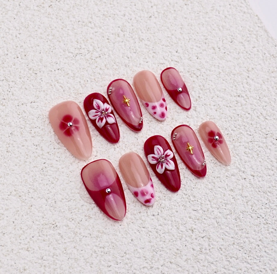 Almond press-on nails featuring cherry red aura blush, white 3D flower, gold star charm, and polka dot french tips.

