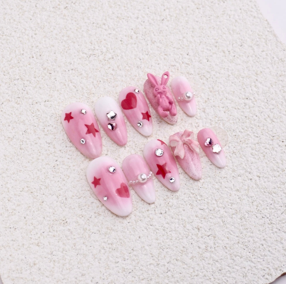 Almond press-on nails featuring pink blush aura, red stars, hearts, 3D textured rabbit charm, and ribbon bow.

