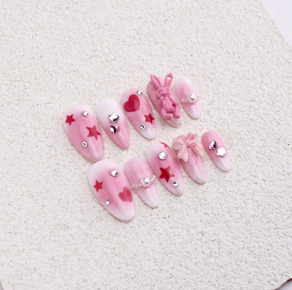 Almond press-on nails featuring pink blush aura, red stars, hearts, 3D textured rabbit charm, and ribbon bow.

