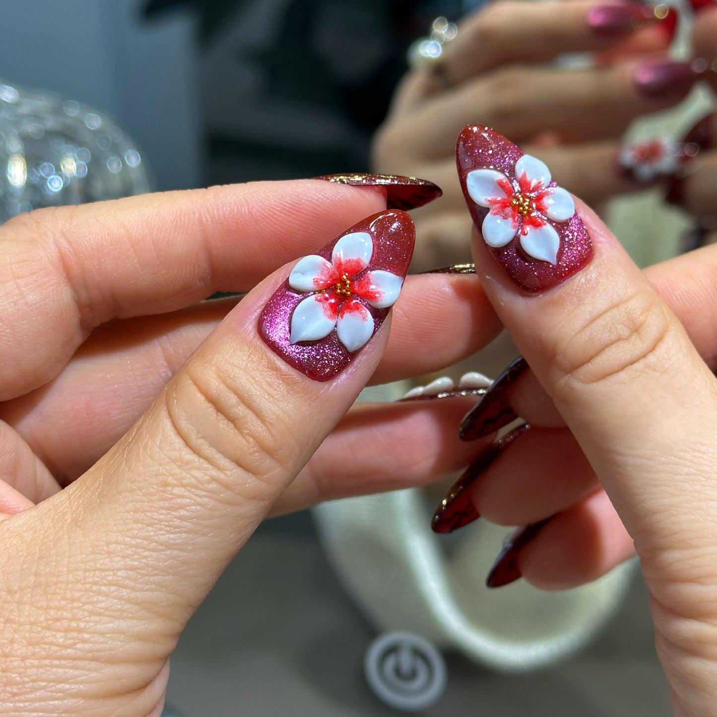 Almond press-on nails featuring wine red velvet cat eye finish, large 3D white flower charms, and gold metallic swirl details.
