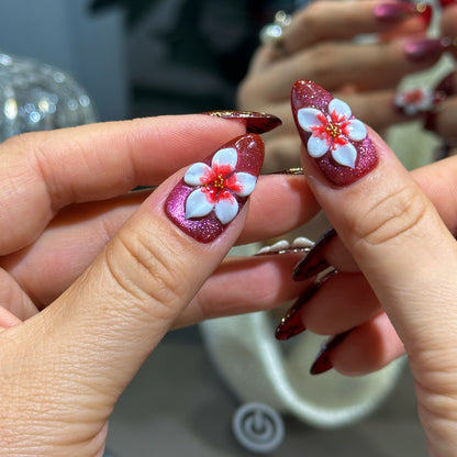 Almond press-on nails featuring wine red velvet cat eye finish, large 3D white flower charms, and gold metallic swirl details.

