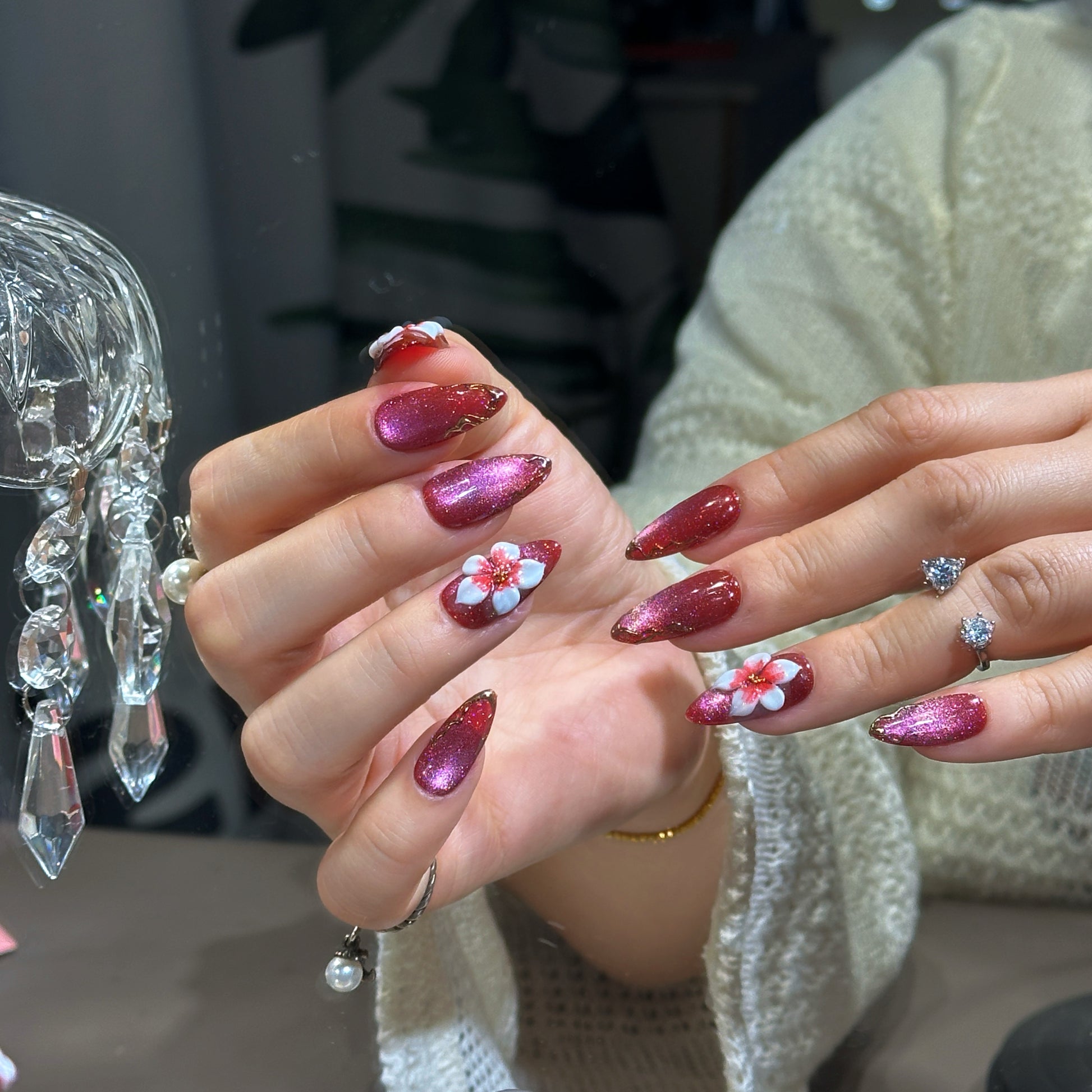 Almond press-on nails featuring wine red velvet cat eye finish, large 3D white flower charms, and gold metallic swirl details.

