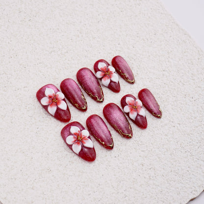 Almond press-on nails featuring wine red velvet cat eye finish, large 3D white flower charms, and gold metallic swirl details.


