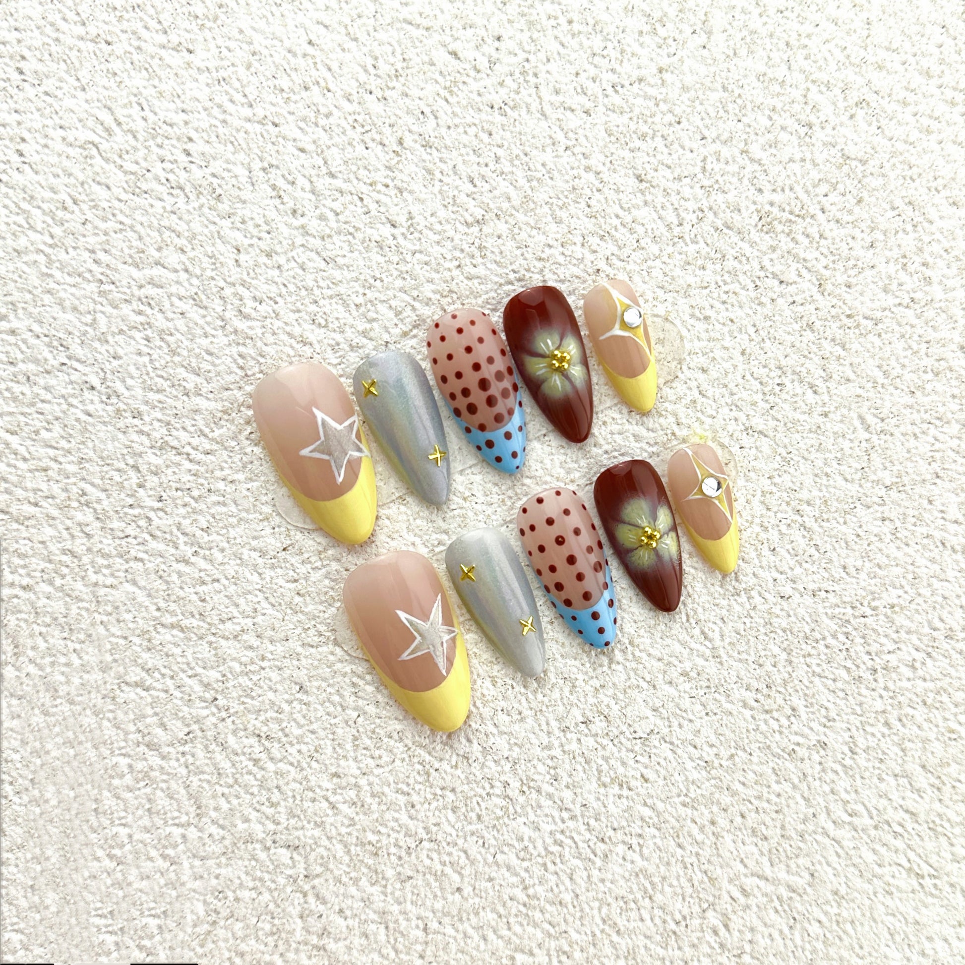 Starry Dots press-on nails flat lay with nude-pink base, yellow French tips, silver chrome, holographic stars and polka dots
