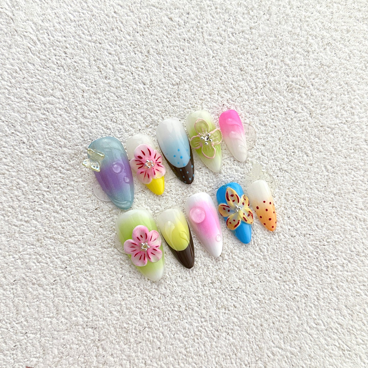 Rainbow Garden press-on nails flat lay with white base, rainbow ombre gradients, 3D flowers, butterfly and polka dot accents
