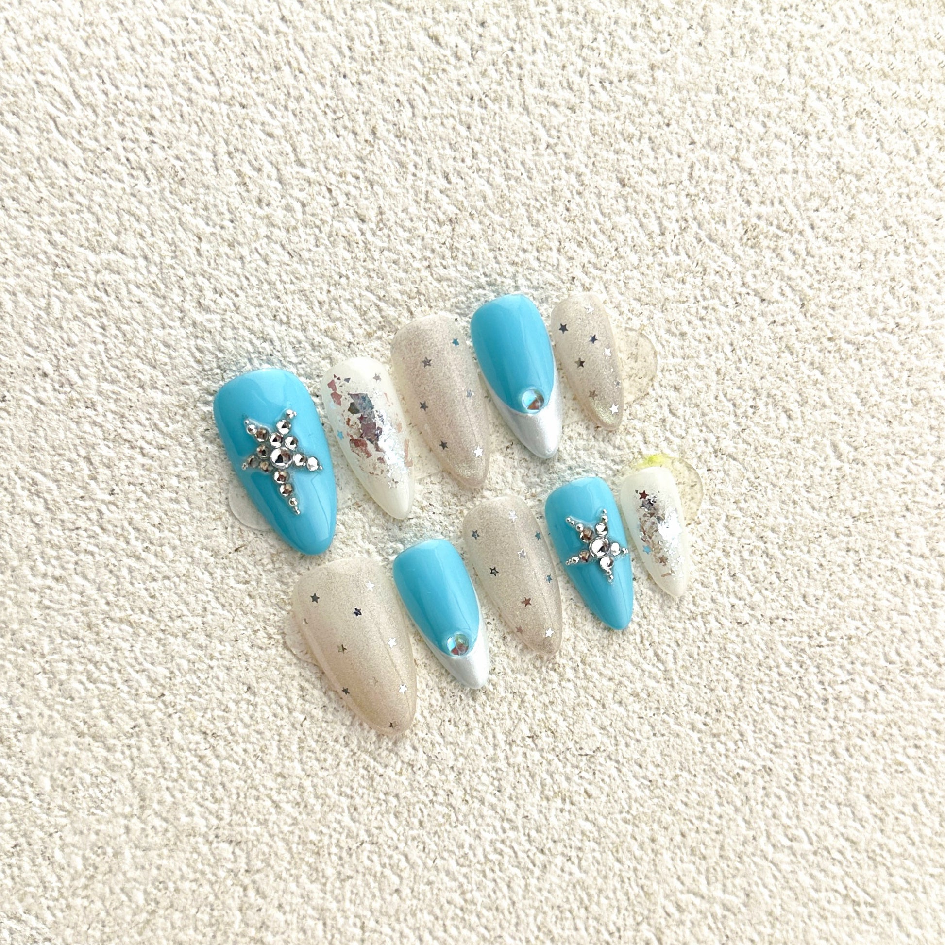 Celestial Cross press-on nails flat lay with sky-blue base, milky white accents, silver glitter and crystal cross embellishments
