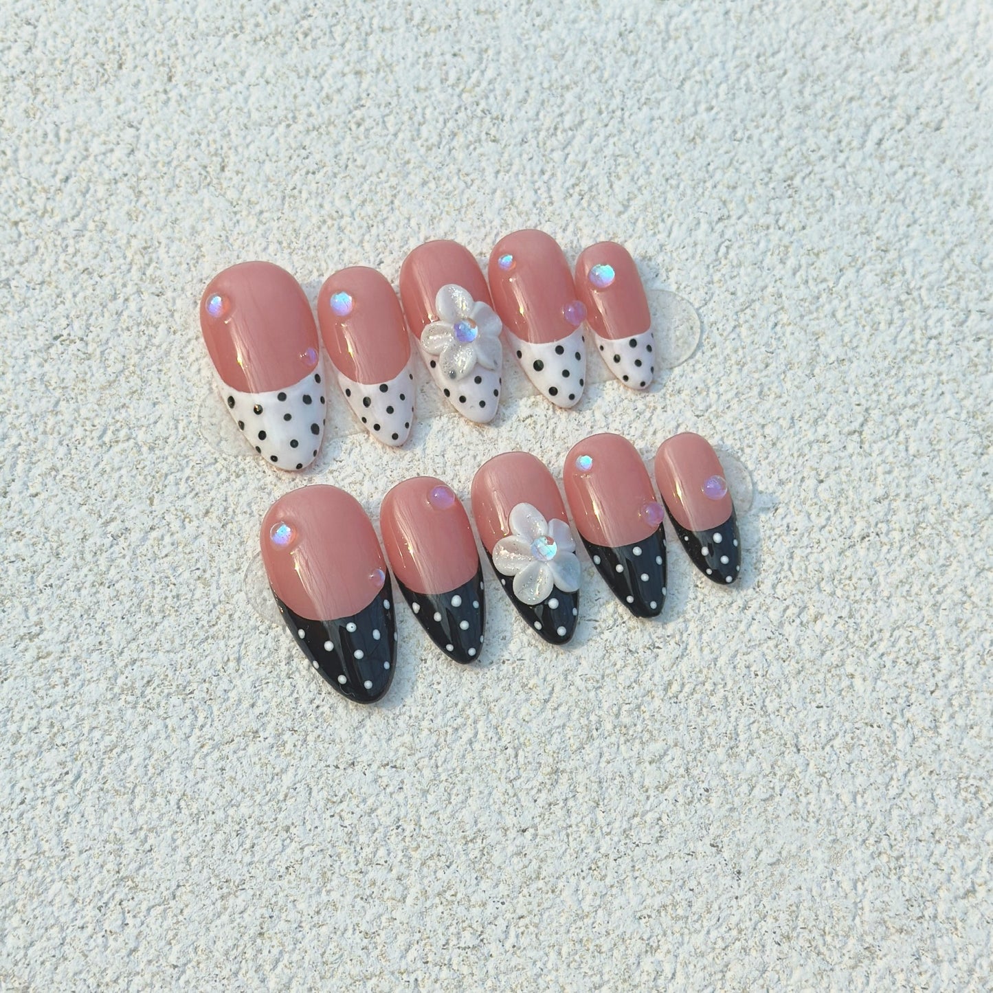 Polka Dot Bloom press-on nails flat lay with nude-pink base, black and white polka dot French tips and 3D white flowers
