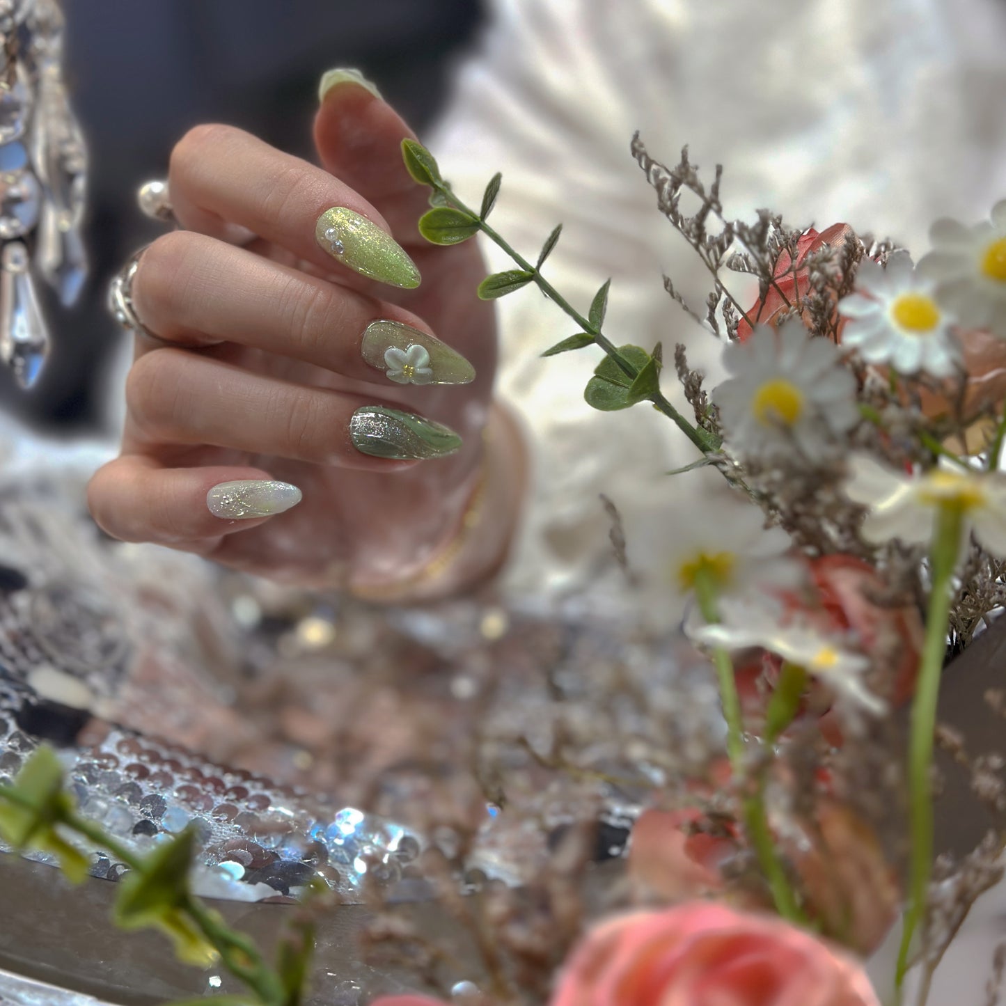 Hand wearing Matcha Daisy Garden press-on nails with roses and daisies showing full-coverage silver glitter on matcha green base

