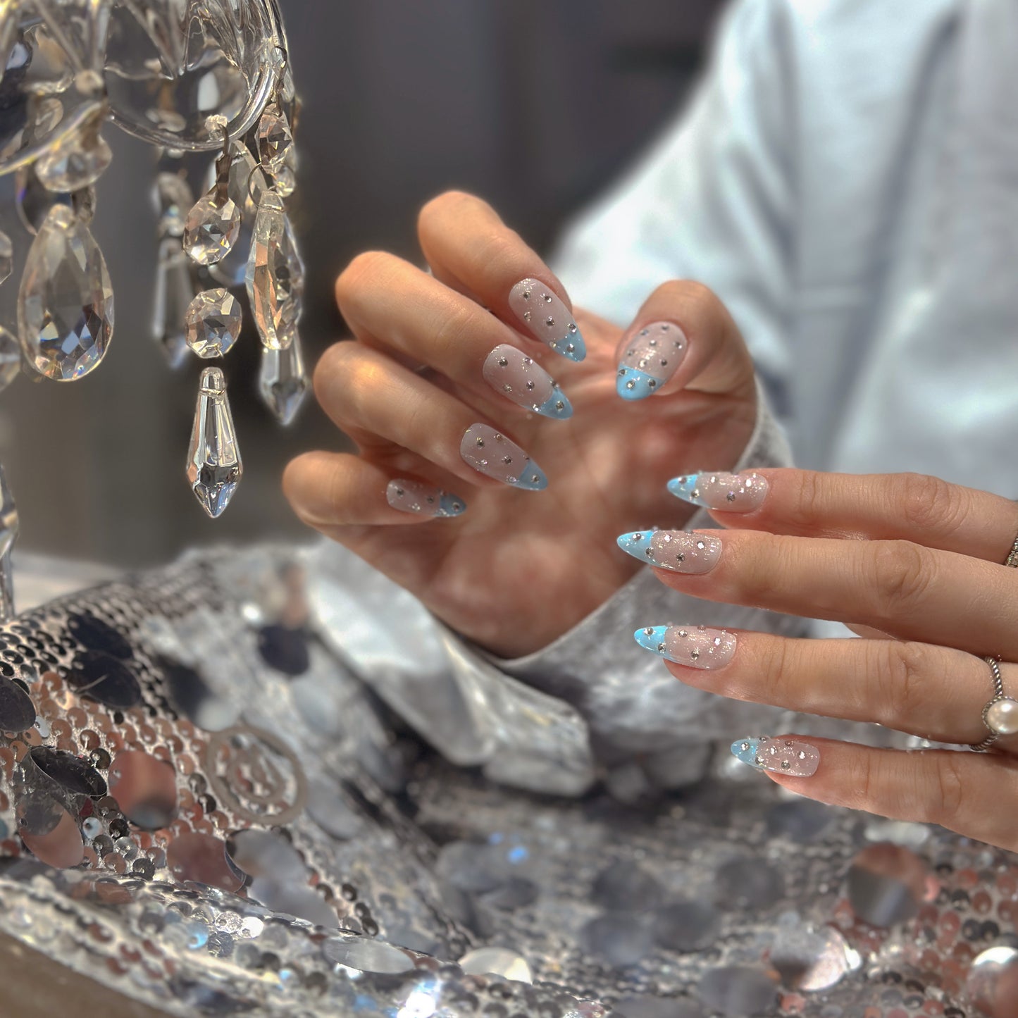 Hand modeling Blue French Crystal Rain nails beside crystal chandelier showing sheer base with mixed-size silver rhinestones
