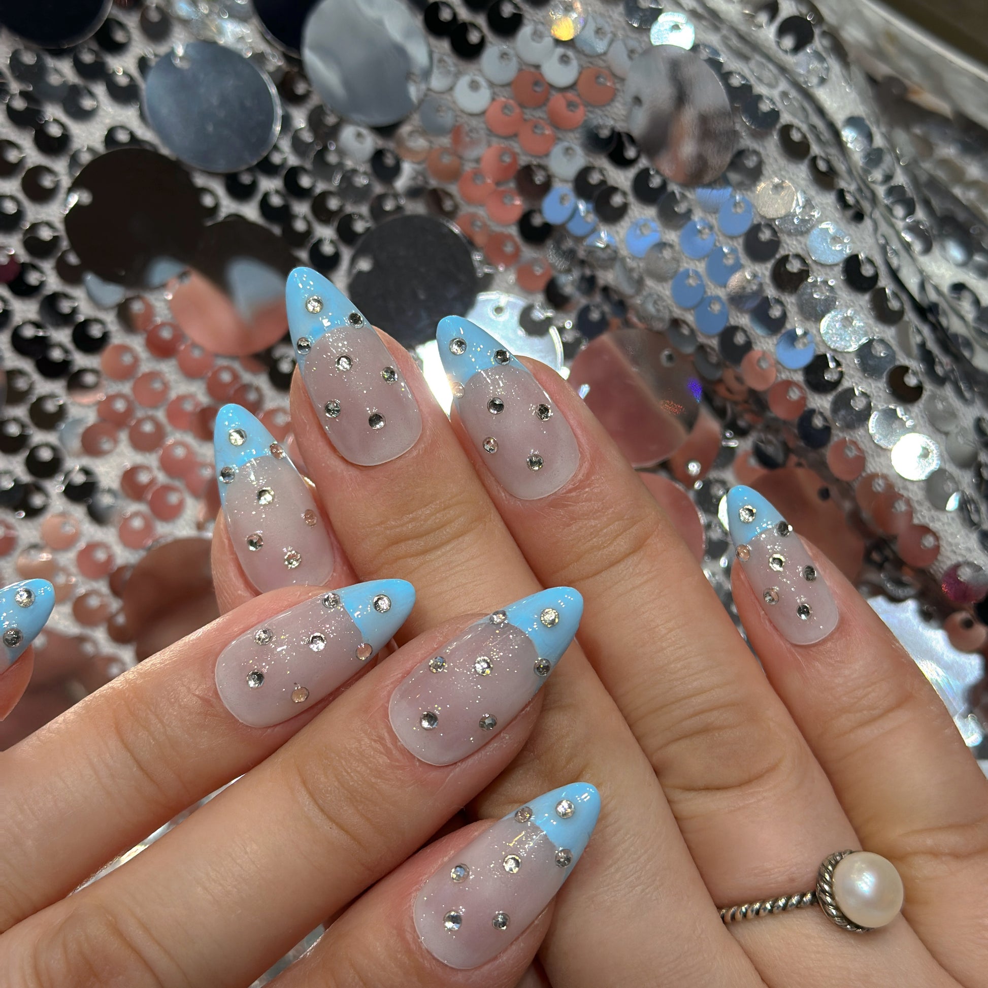 Both hands with Blue French Crystal Rain nails against silver sequin background displaying scattered rhinestones and glass-like transparency

